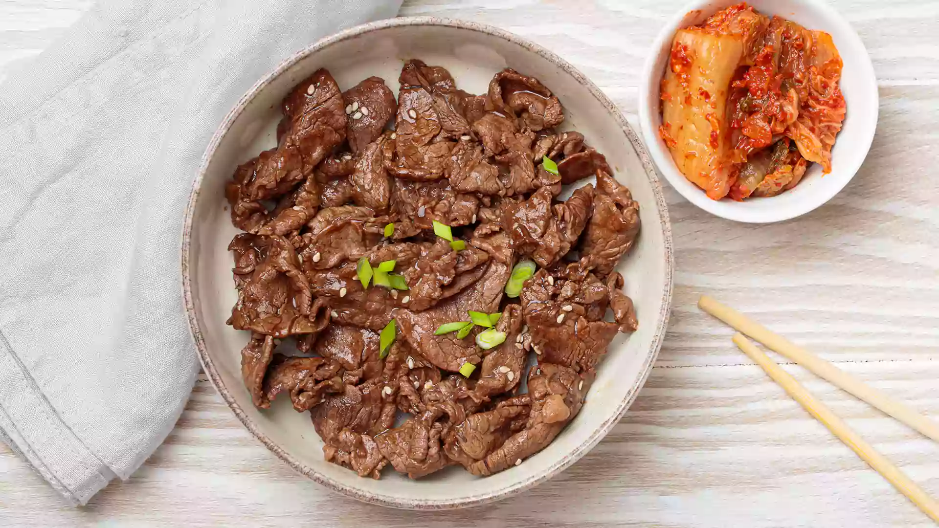 Sepiring bulgogi daging sapi yang dimasak dengan bumbu manis gurih, dihiasi wijen dan daun bawang, disajikan bersama kimchi dalam mangkuk kecil.