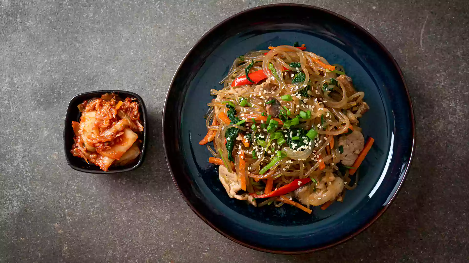 Hidangan japchae mi bening khas Korea dengan sayuran warna-warni dan daging, ditaburi wijen, disajikan bersama kimchi dalam piring kecil.