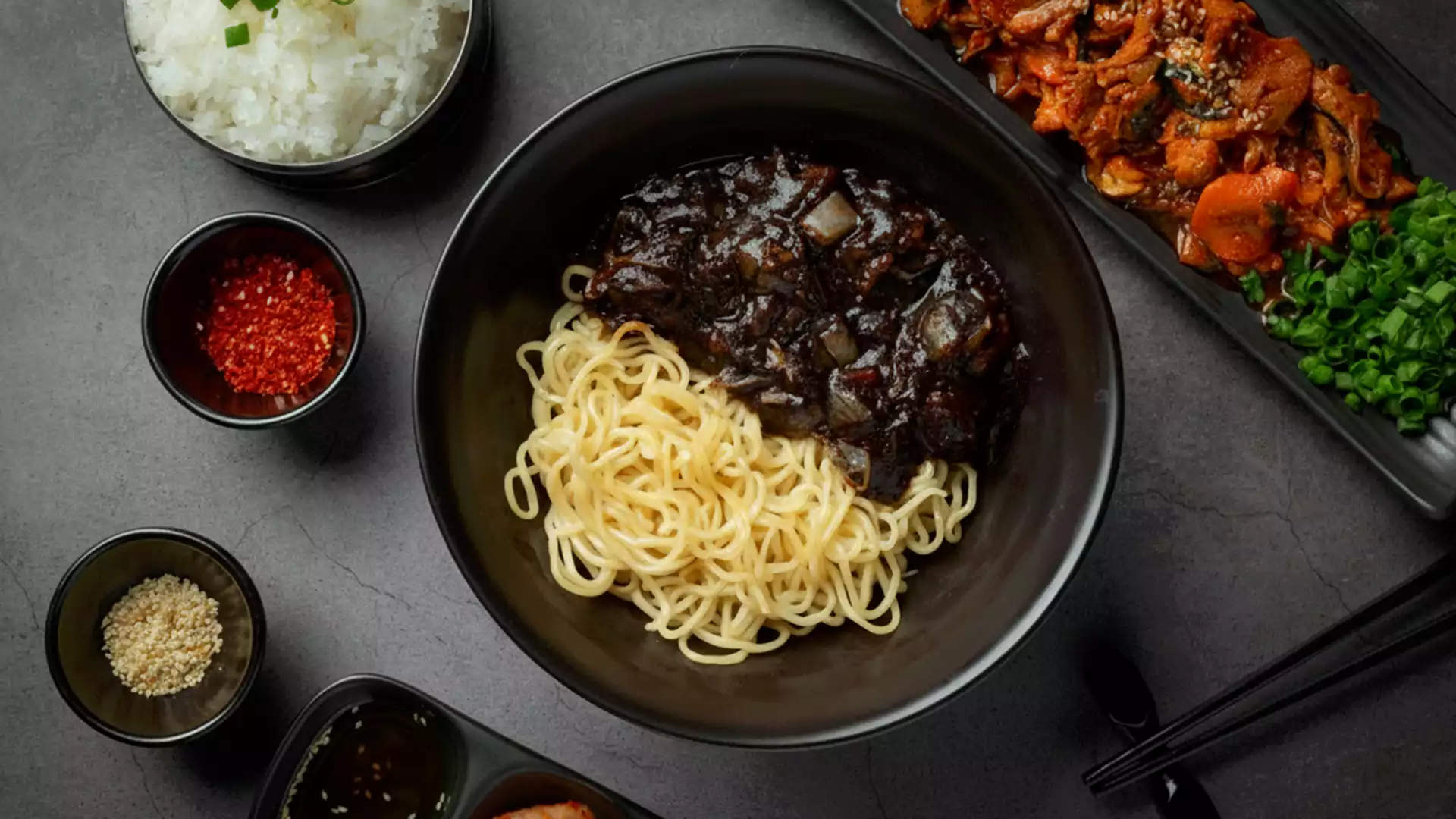 Semangkuk jjajangmyeon dengan mi kuning dan saus kedelai hitam khas Korea, disajikan bersama lauk dan bumbu pelengkap di atas meja hitam.